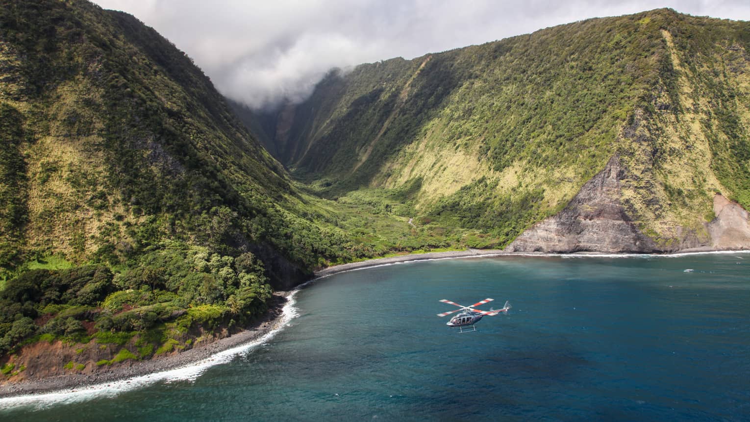 Helicopter flies over ocean by sweeping green volcanic mountains