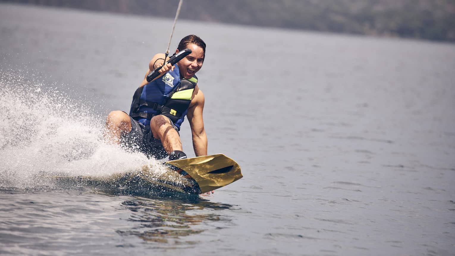 Young man wearing a blue and green life vest and blue swim trunks wakeboarding through the water