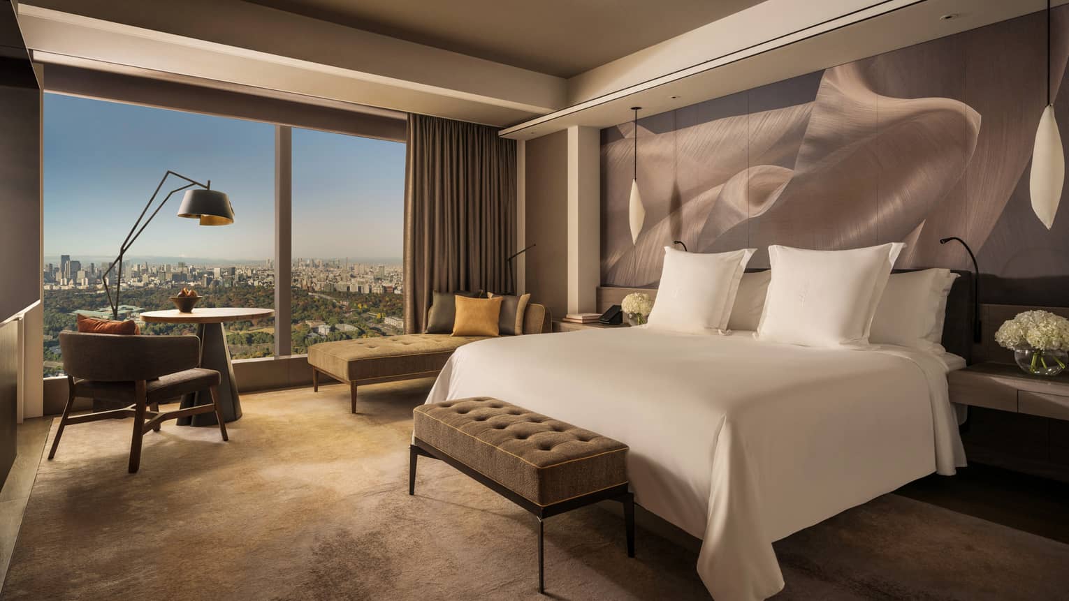Hotel room with a cushioned bench at the end of white king bed and a wall of windows