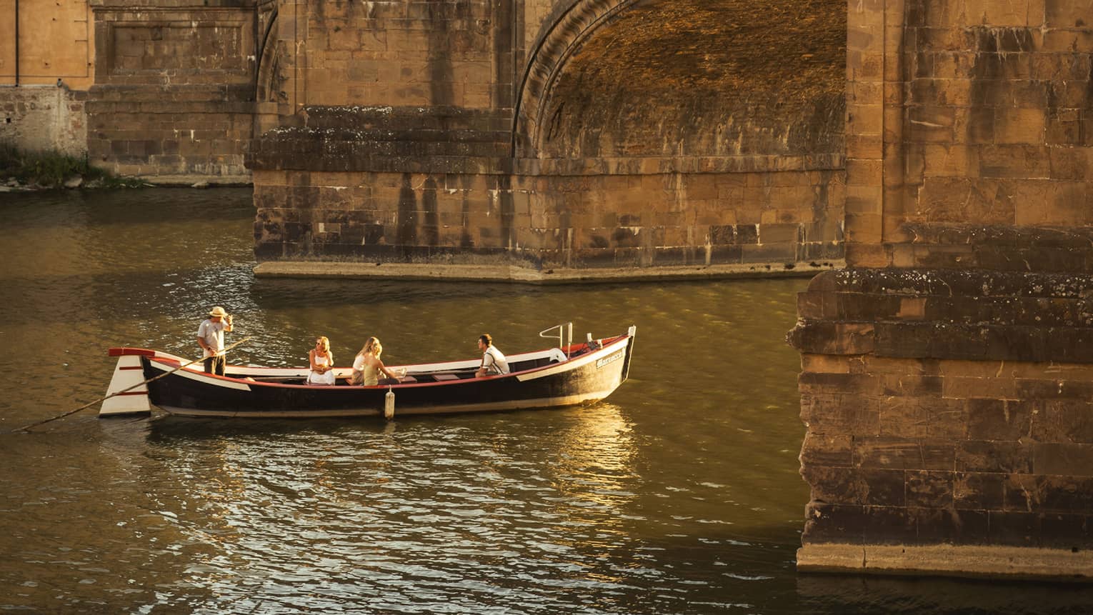A barchetto, an ancient boat, with a few people on it are leisurely making their way under a crowded stone bridge.