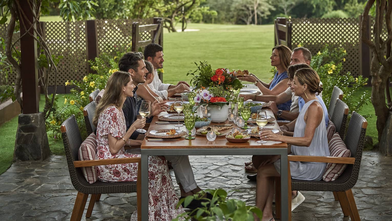 Eight people sit around a long dining table set up outside beneath a canopy of greenery and string lights