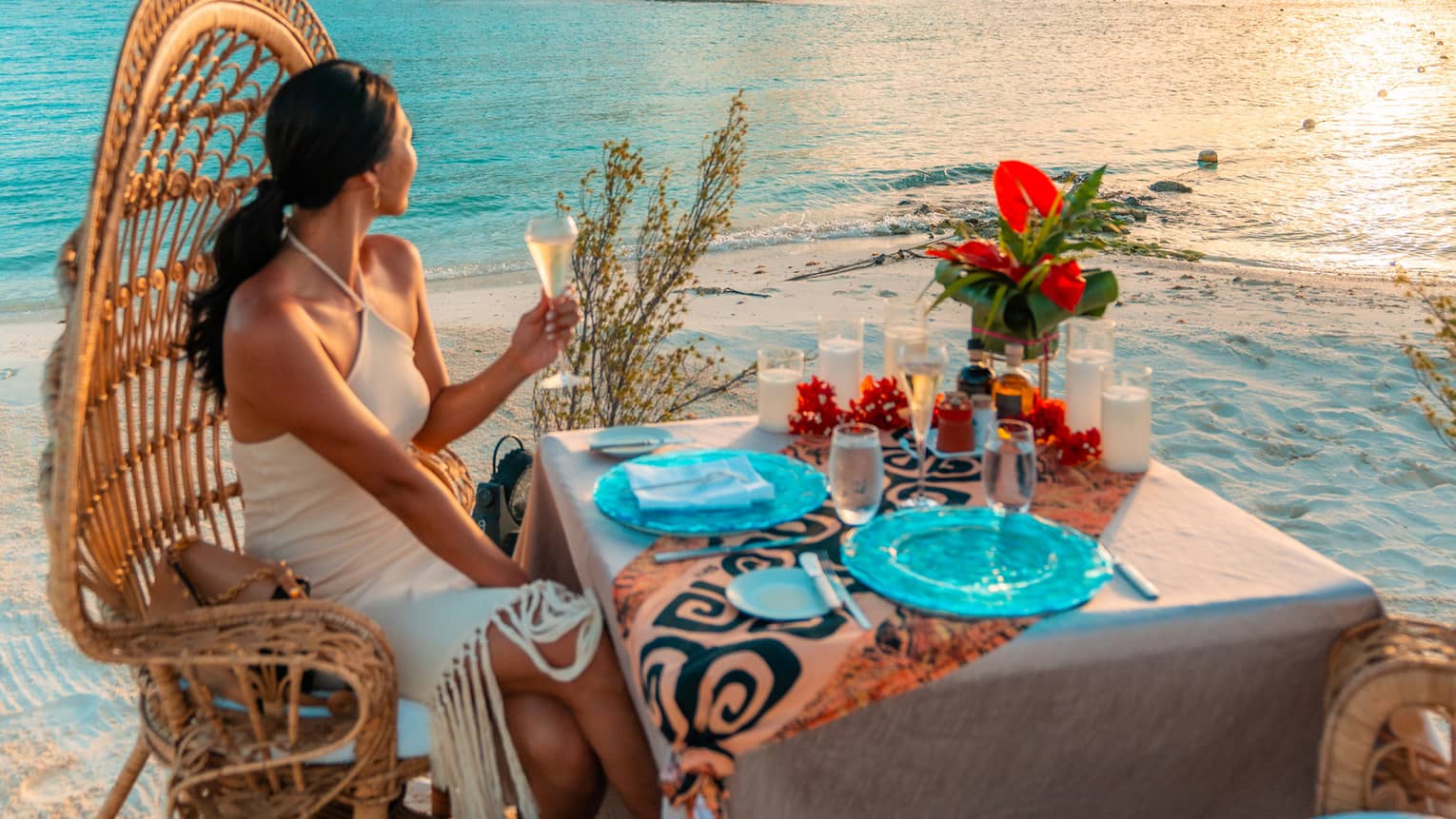 Person in high-backed wicker chair sits at table on beach, holding glass of white wine and gazing at the sunset