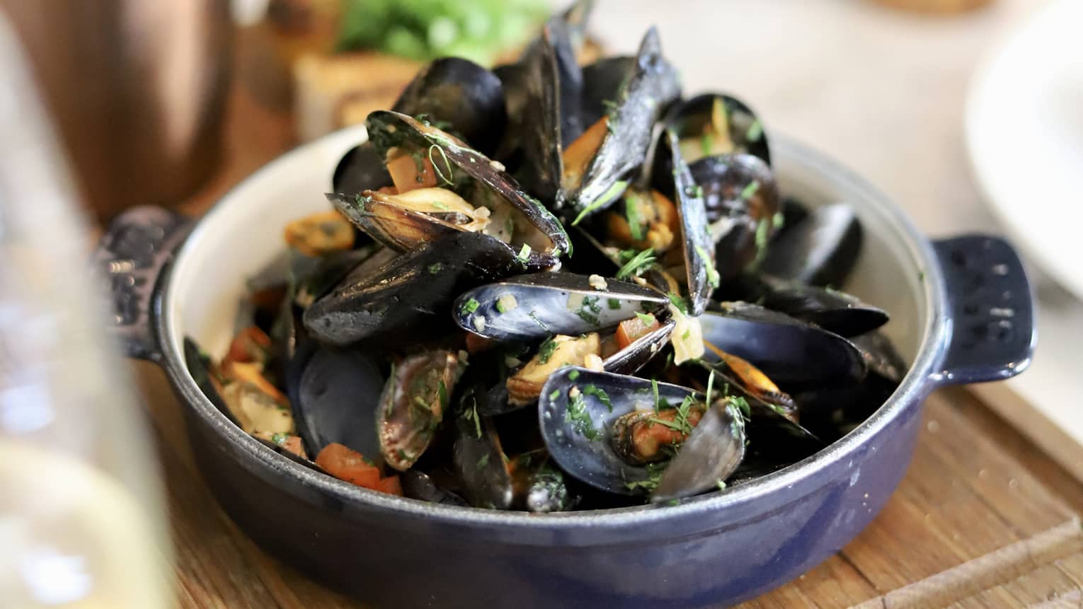 Mussels served in a dark-blue porcelain pot set atop a wooden serving platter with a side of French fries in the background