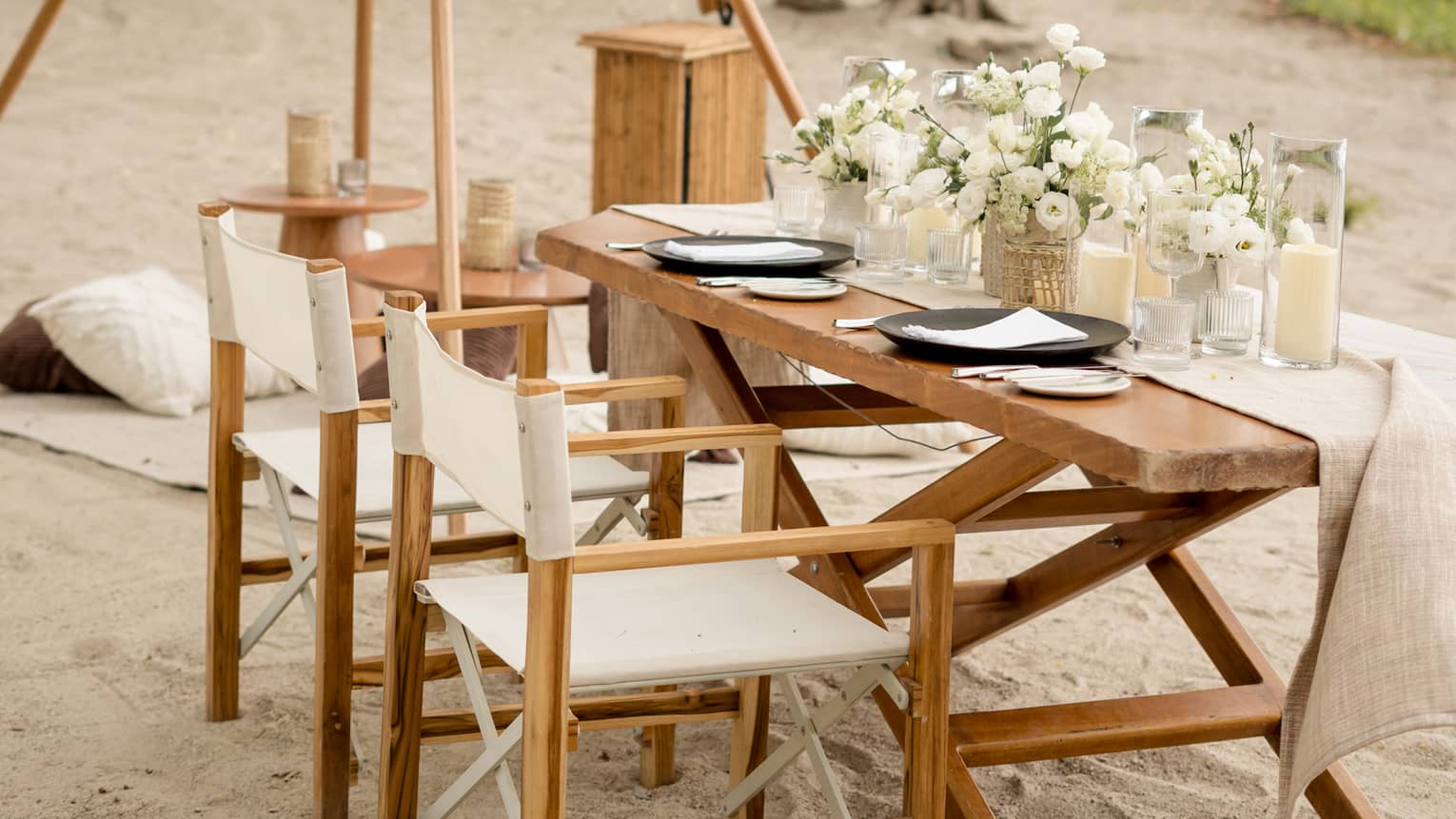 Wooden dining table and chairs set up on a beach, decorated with white floral centrepieces and candles