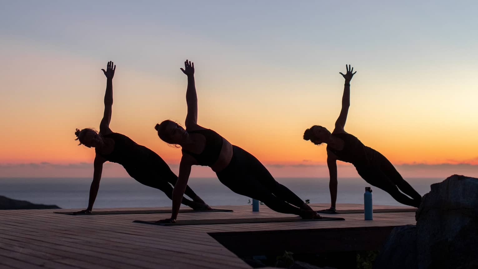 Three guests doing side plank poses on yoga mats on a wooden dock are silhouetted against a clear blue, orange and pink sky.