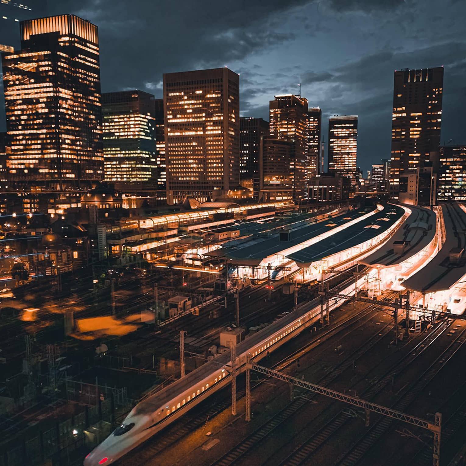 High-speed train passing through a city railway station at night with illuminated skyscrapers in the background