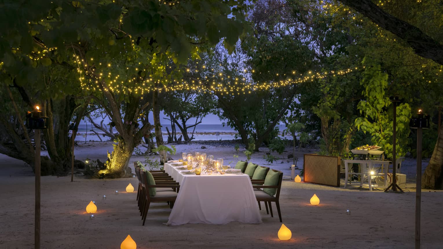 Long dining table, chairs with cushions on sand, surrounded by candles and strings of lights