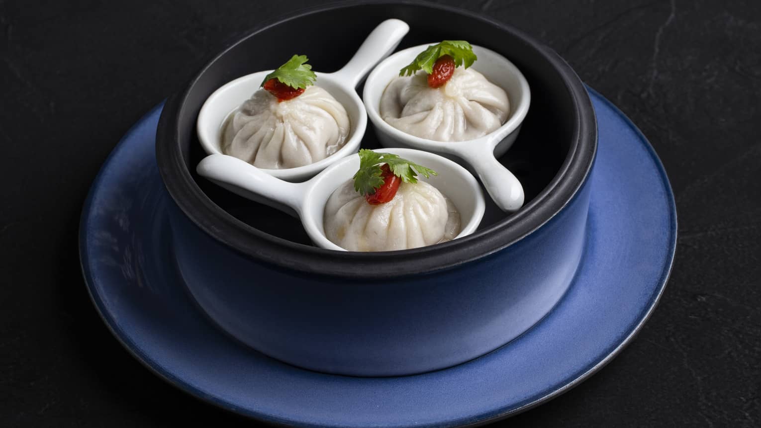 Three dumplings with garnish on white spoons in a blue bowl with a matching plate.