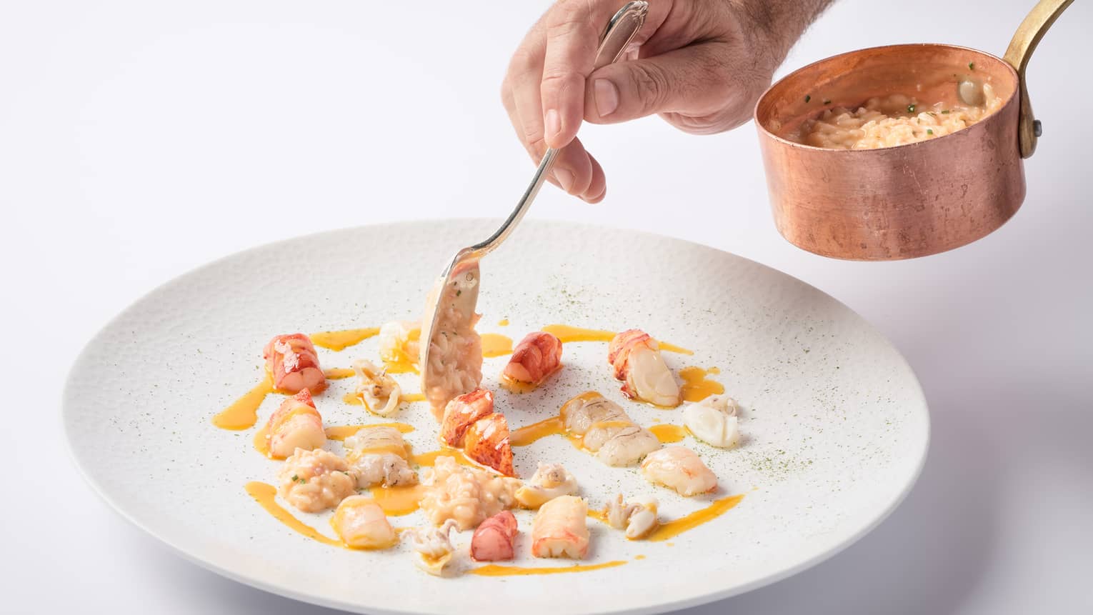 A close up of a hand spooning out a cream sauce and vegetables from a copper ladle onto a porcelain plate.