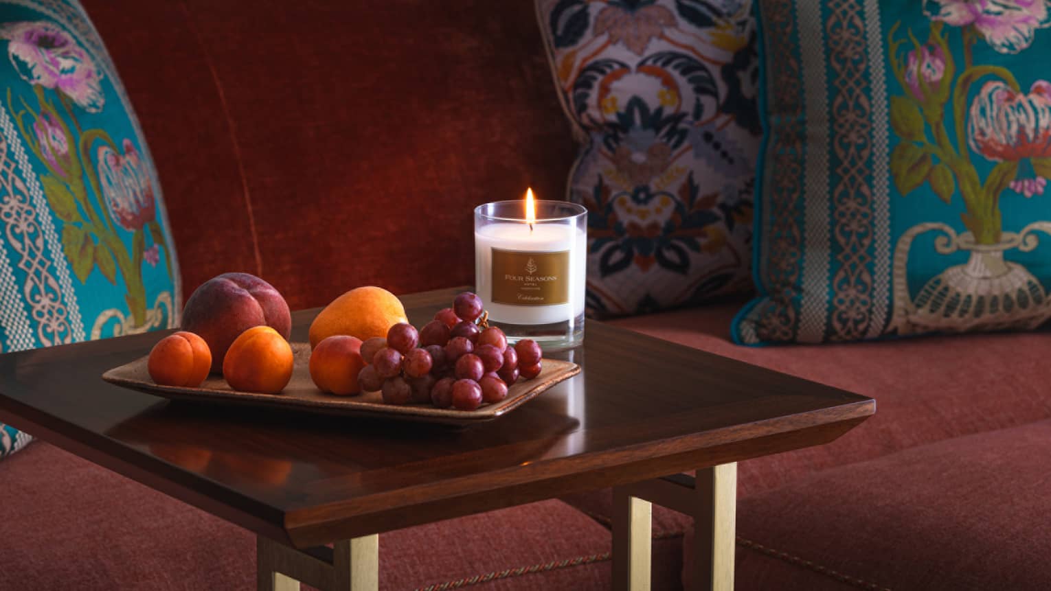Couch tray with fresh fruit and Four Seasons lit candle, red loveseat with patterned pillows in the background