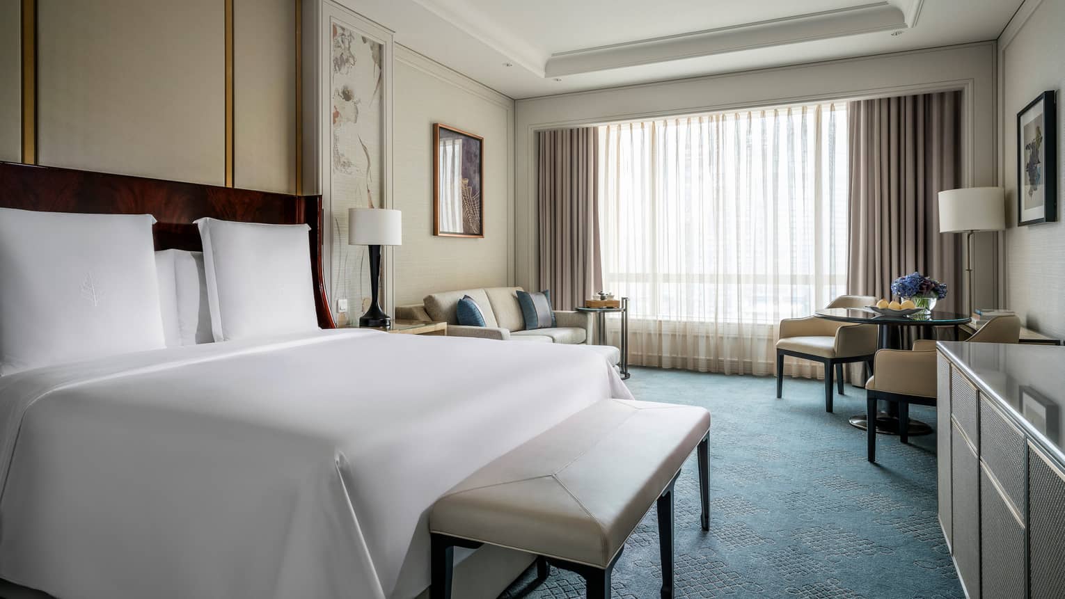 Deluxe Room bed with white linens, bench at foot, sofa and cafe table by window