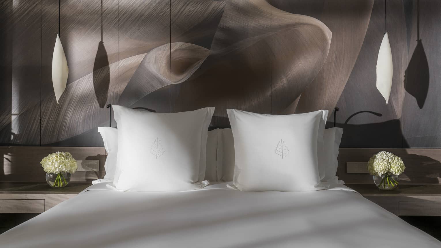 Close-up of a Four Seasons king bed with white pillows and soft lighting, flanked by bedside tables holding white flower arrangements