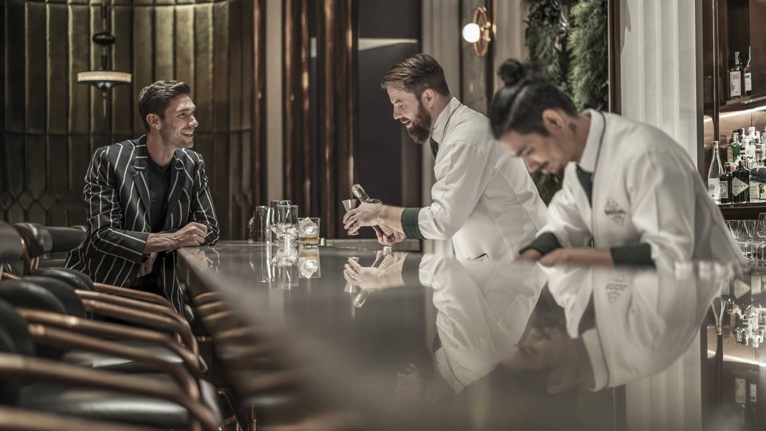 Bartender preparing a drink while a customer sits at the bar, both smiling, in a modern, upscale bar with a reflective countertop and warm lighting.