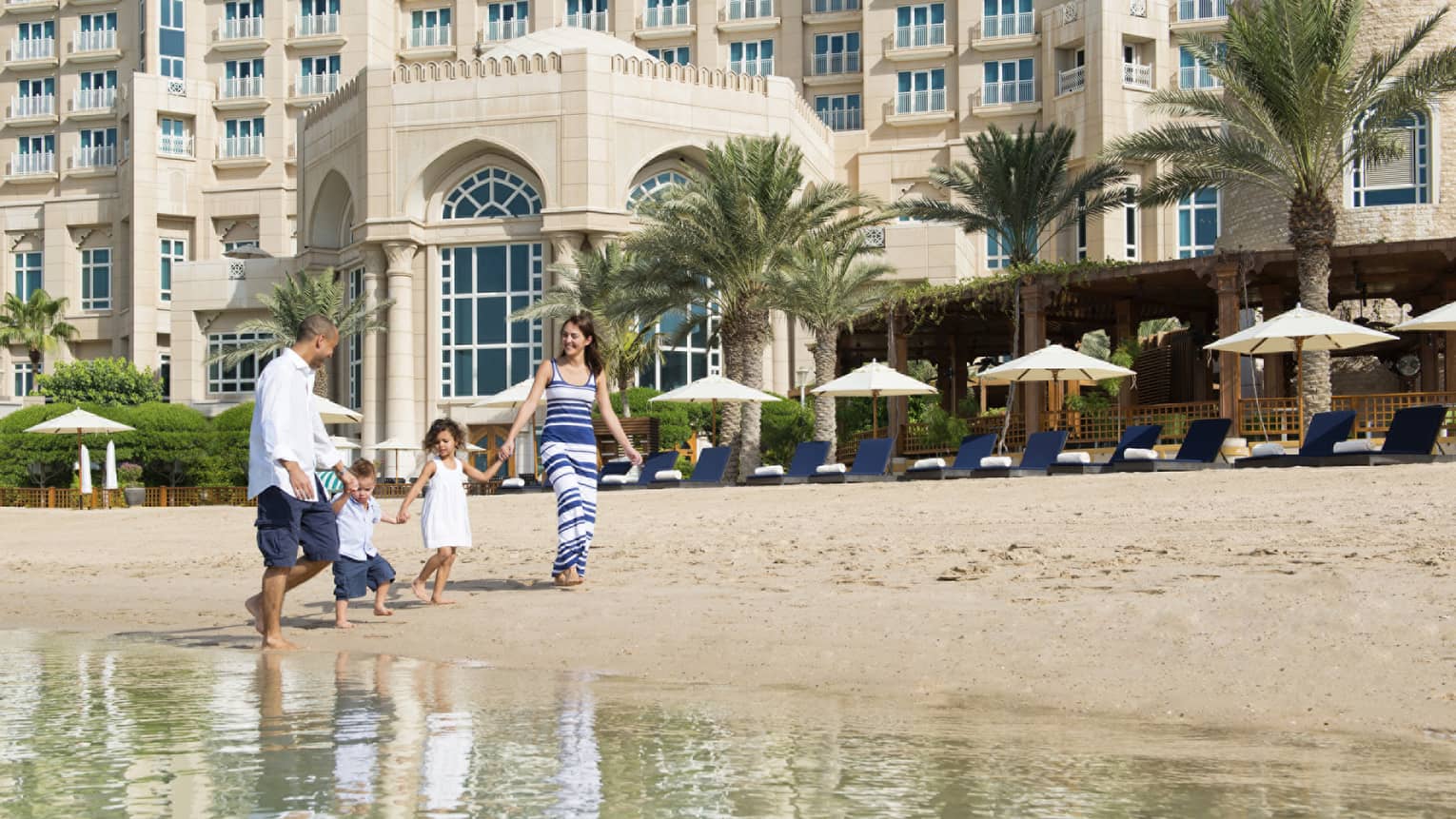 Woman and man hold hands of two young children, walk barefoot in sand along beachfront
