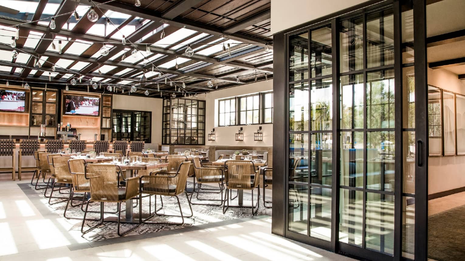 Dining room and bar with glass roof, modern wicker chairs and tables, glass sliding doors with black frames, glass bulb light strings