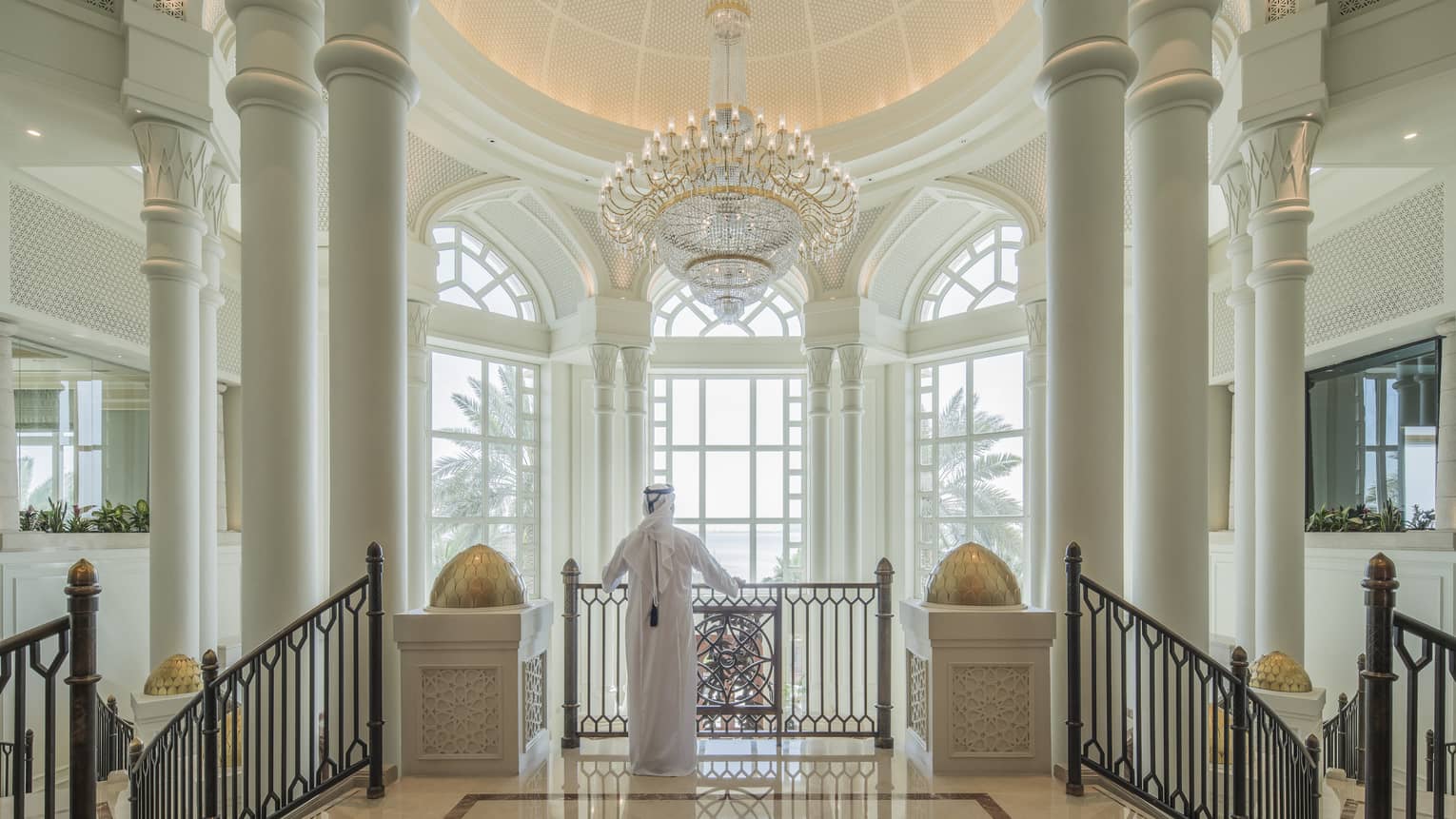 A man in a white thobe in front of enormous floor-to-ceiling windows in a marble foyer. 