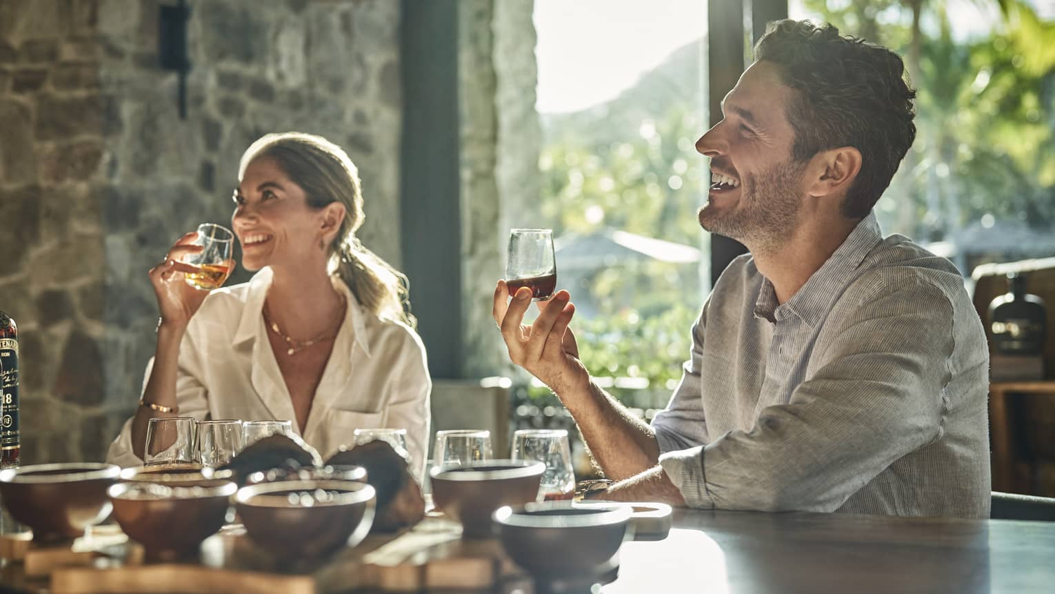 A man and a woman sit at a counter looking up and smiling and each holding a glass of rum with various other glasses and ingredients sit in front of them on the counter