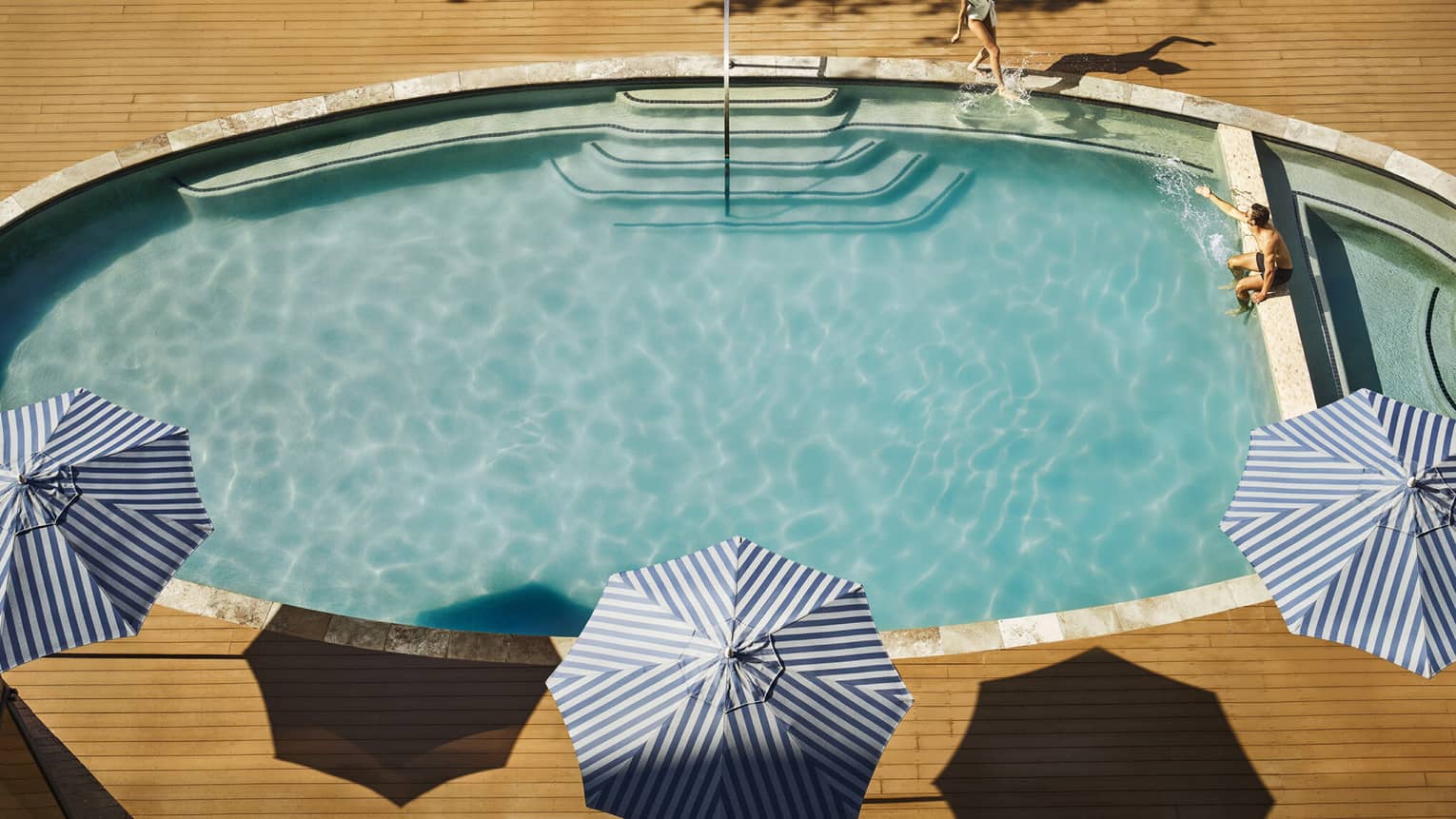 Aerial view of a couple lounging at an oval-shaped pool lined with blue striped umbrellas