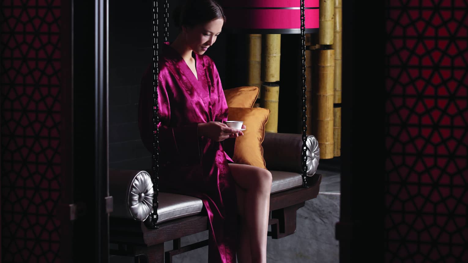 Woman in red silk robe sits on bench with small teacup in dark spa