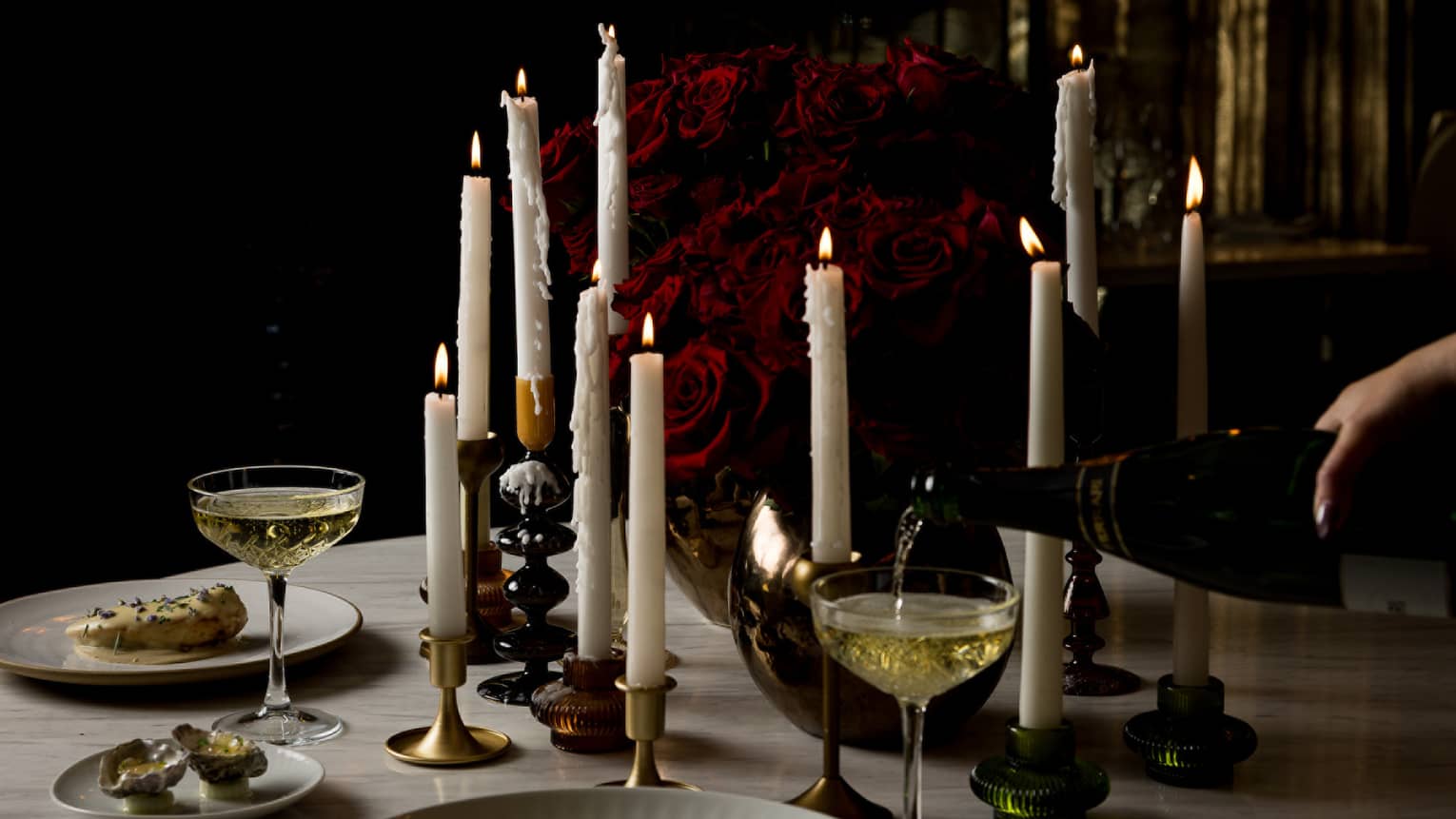 A round marble table topped with various pillar candles, a red rose centrepiece and food and drink for two is set in a dimly lit room