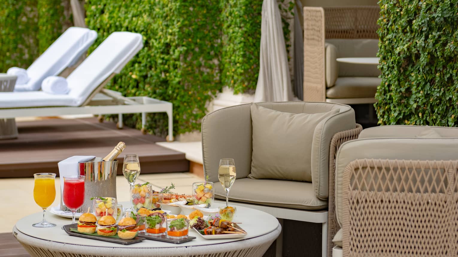 A lush cabana area under an umbrella with chairs and loungers, with an alluring array of appetizers and drinks on a low table.