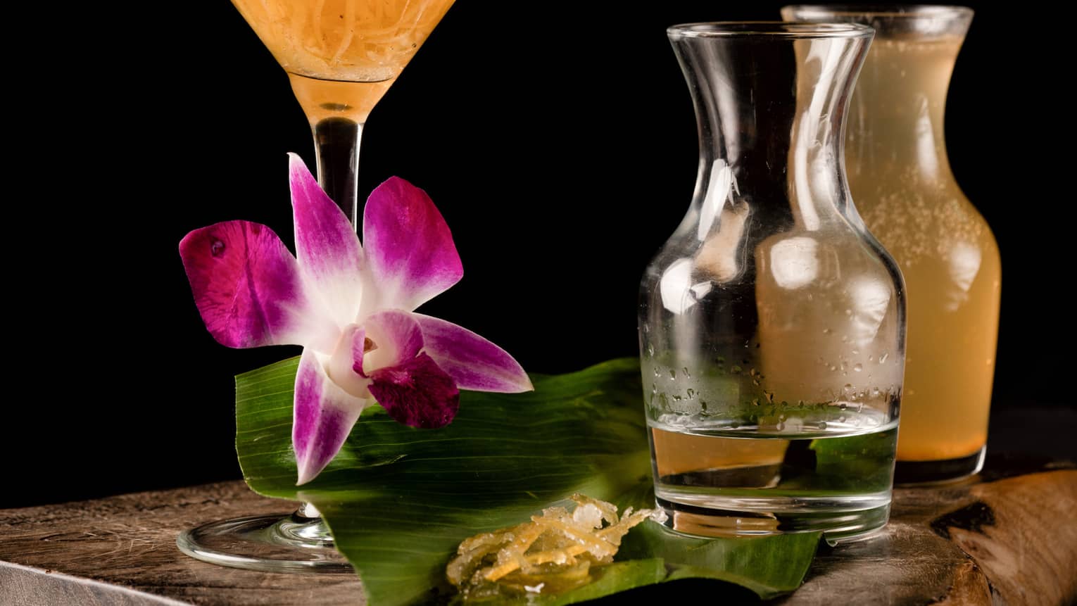An orange martini glass on a wooden table surrounded by several pitchers of alcohol.