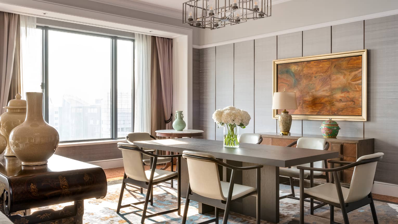 A sophisticated dining room with a rectangular wooden table, cream-coloured chairs, decorative vases and large window. A framed painting and lamp adorn the sideboard.