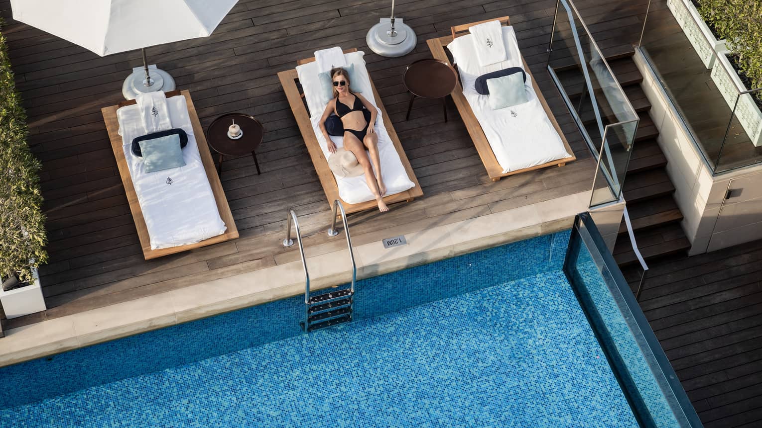 Aerial view of person wearing black bikini and sunglasses lounging on a poolside daybed on a rooftop deck