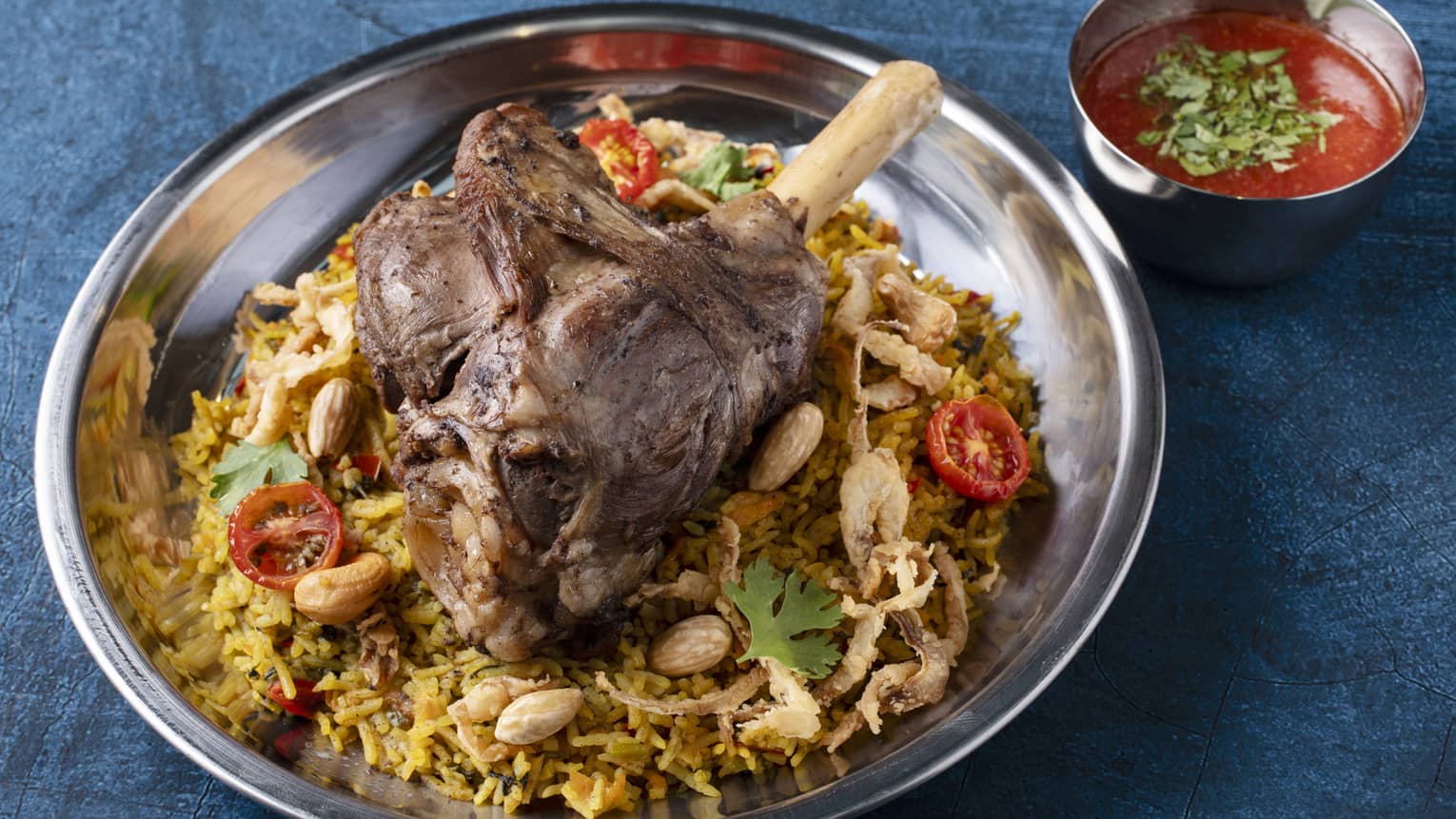 Traditional kabsa basmati rice with braised lamb shank, nuts, crispy onions and a small bowl of tomato sauce on the side