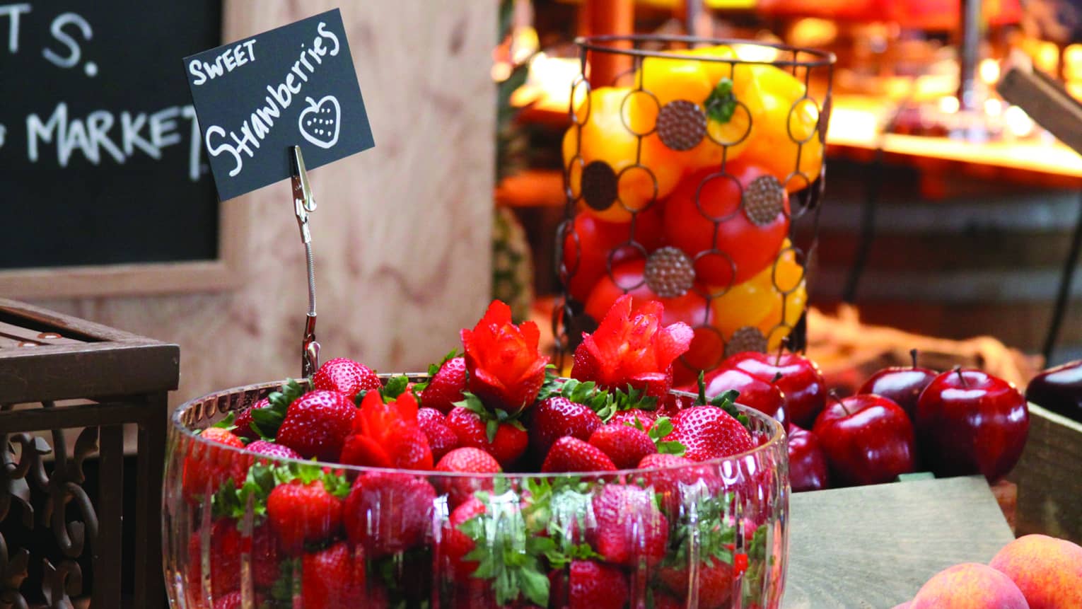 A table in a market displaying a glass bowl of fresh strawberries and an assortment of apples, peaches and peppers.