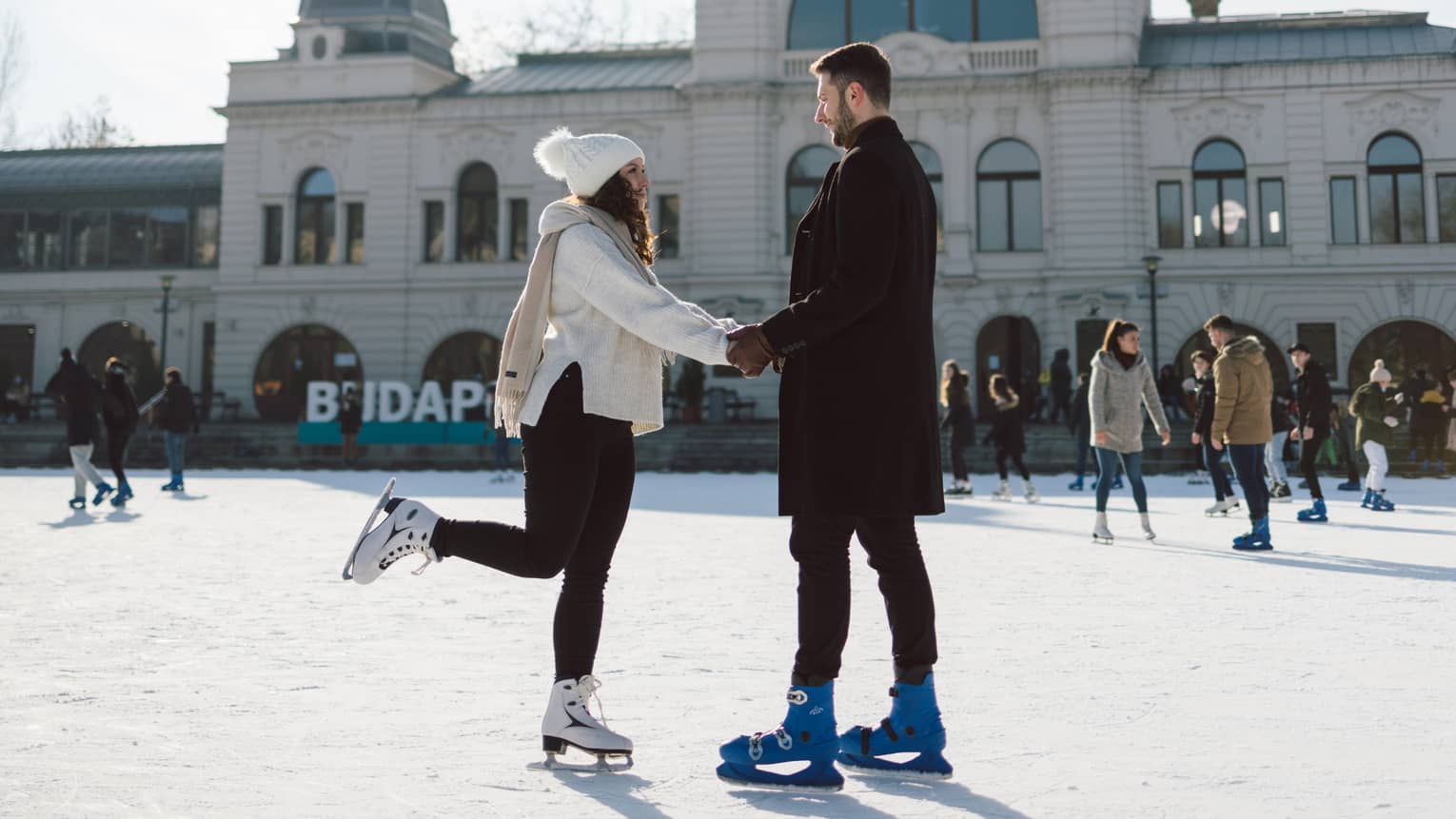 Couple dressed in warm clothing, holding hands while ice-skating on city rink