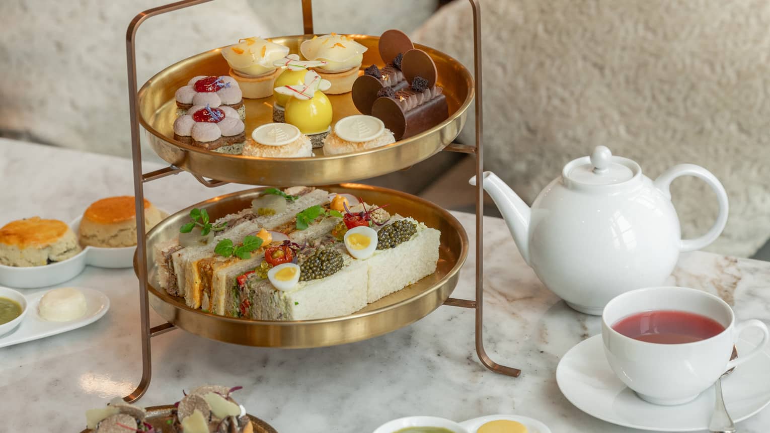 Two-tiered afternoon tea spread on marble coffee table in front of tan sofa