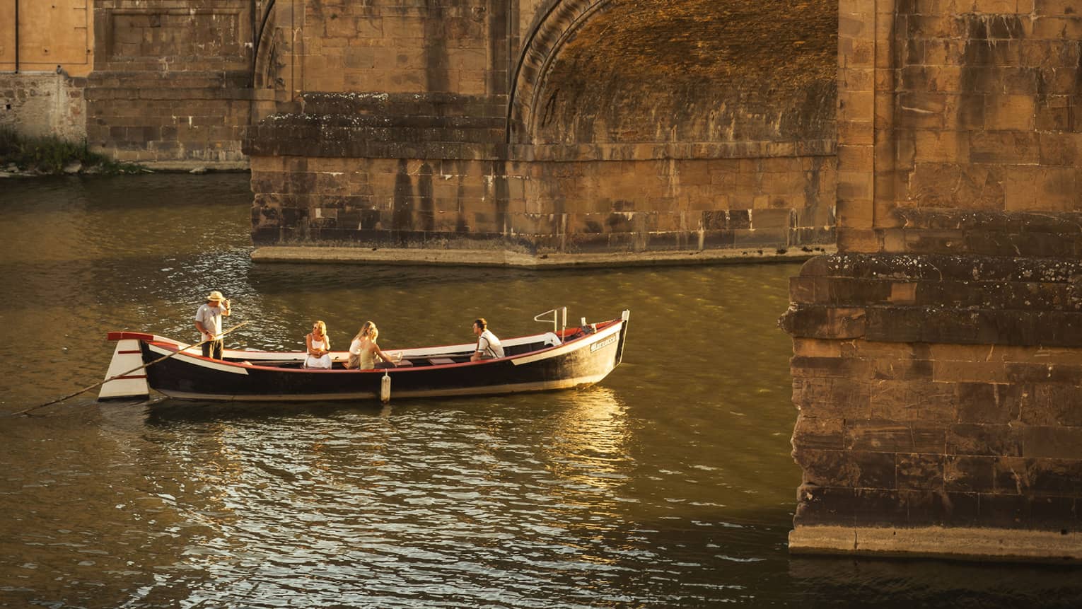 A barchetto, an ancient boat, with a few people on it are leisurely making their way under a crowded stone bridge.