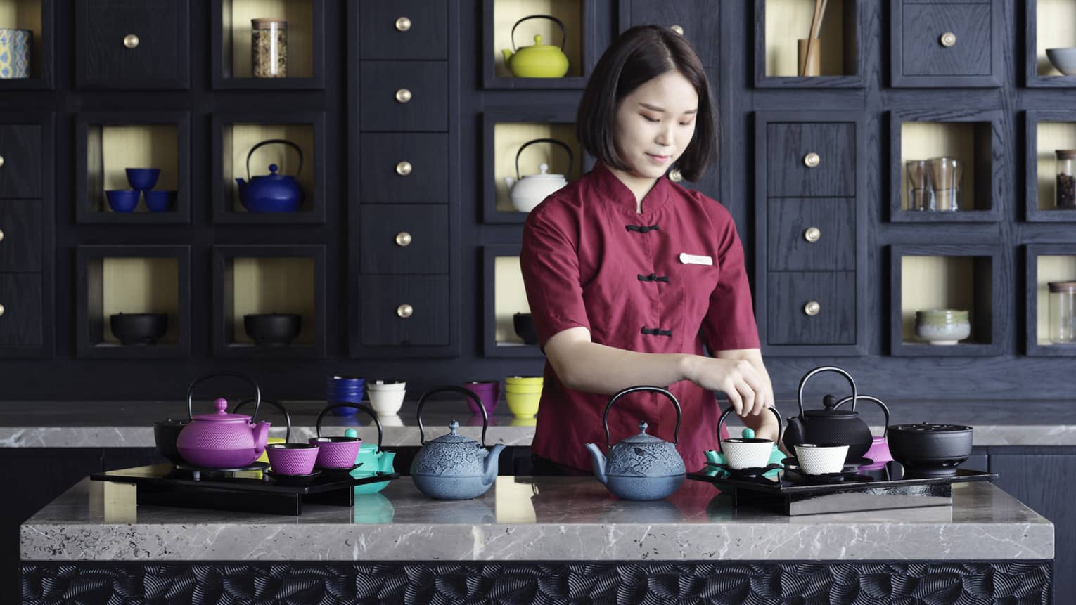 Woman in burgundy uniform prepares colourful iron teapots on marble counter
