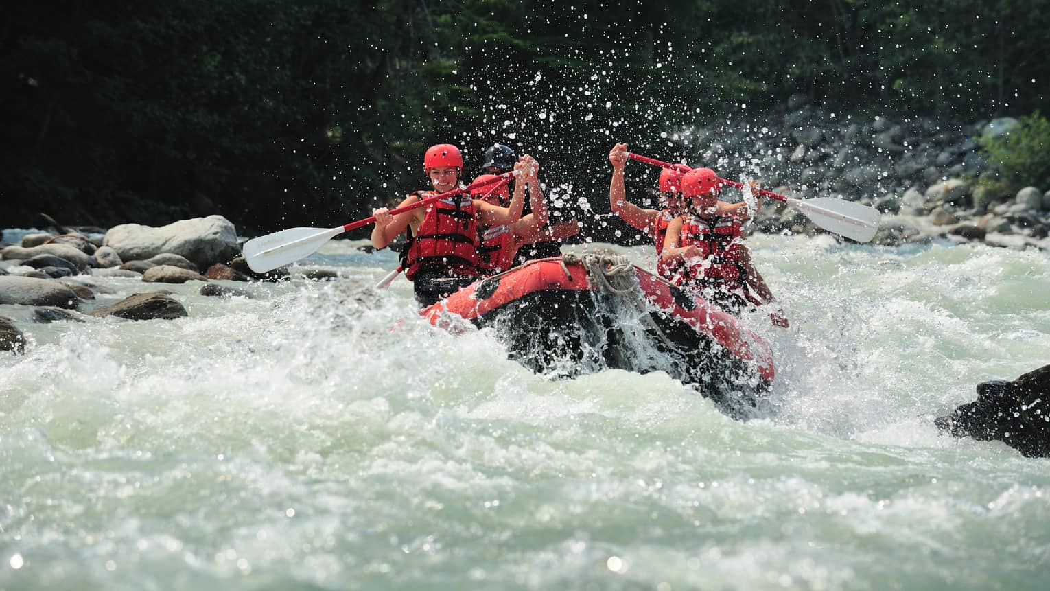 Smiling paddlers in red helmets and lifejackets steer an inflated raft through raging white water, passing evergreen trees.