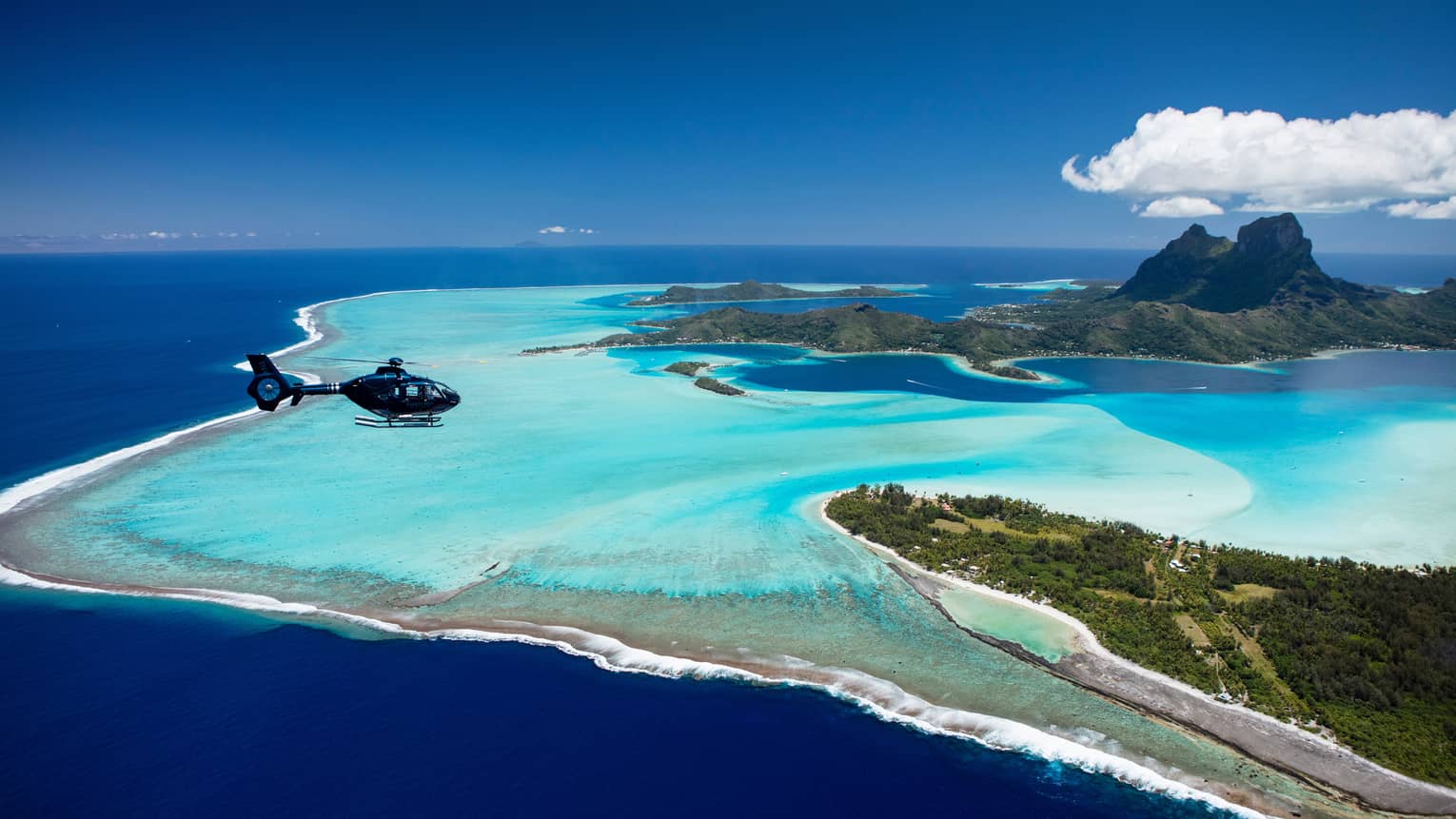 Aerial view of a helicopter flying over Bora Bora and the distinctive pastel blue lagoon that surrounds the islands.