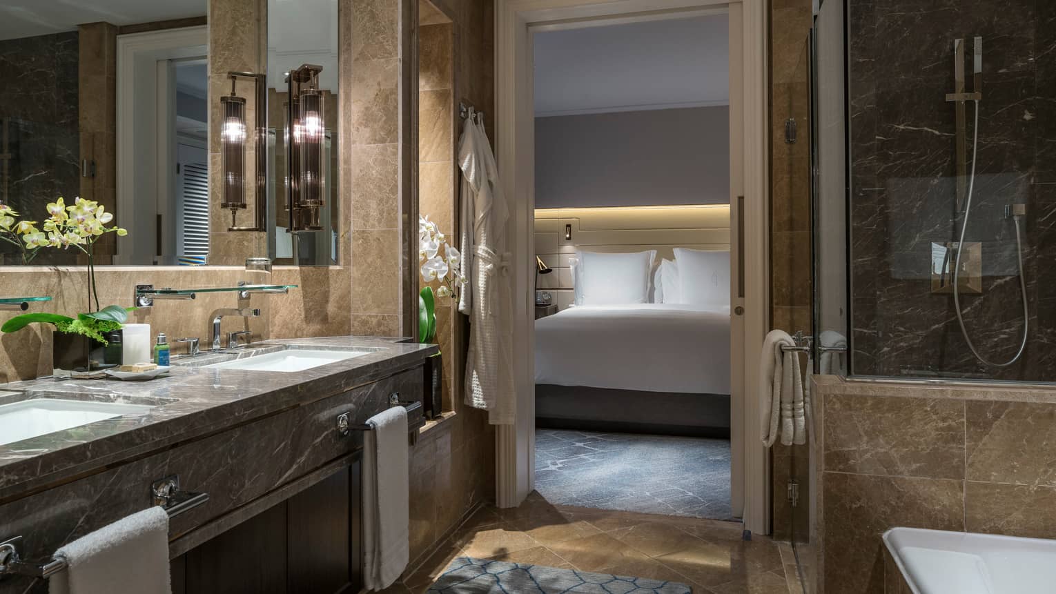 A marble bathroom with double sinks, glass-enclosed shower and bathtub. The bedroom, featuring a well-lit bed with white linens, is visible through a sliding door.