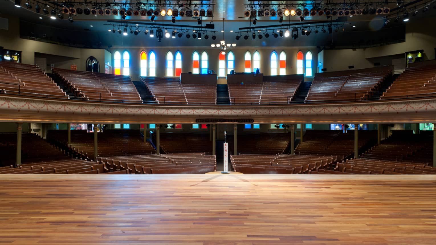 An auditorium interior from the vantage point of a wood-floored stage. The auditorium has balcony-level seating and large stained glass windows behind the seating area.