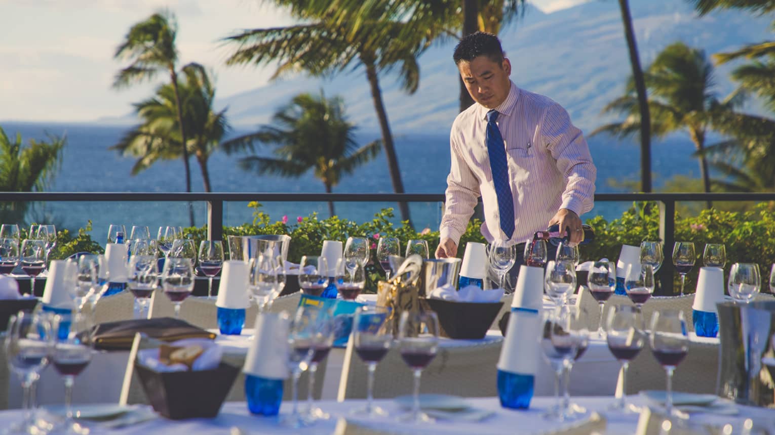 Four Seasons staff pours red wine into glass on table outdoors, surrounded by palm trees, ocean and mountains