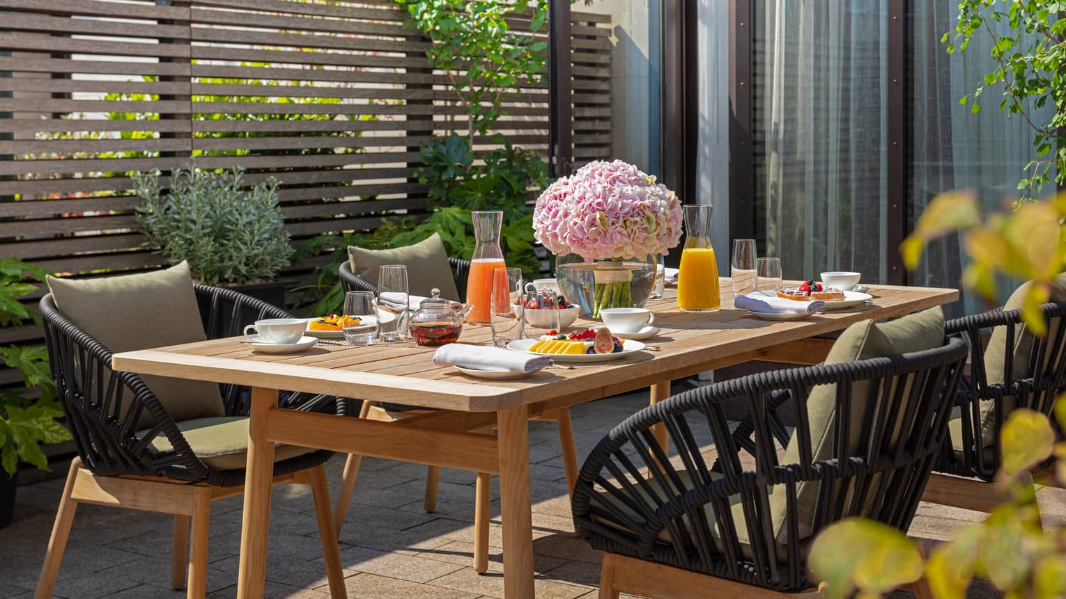 The Executive Conservatory at Four Seasons London Park Lane in Mayfair features an inviting outdoor dining area. A rustic wooden table is set with a colorful breakfast spread, including fresh fruits, pastries, juices, and tea. The table is beautifully decorated with a large bouquet of pink hydrangeas, enhancing the fresh, vibrant setting. Stylish black wicker chairs with comfortable cushions surround the table, nestled amidst lush greenery and a modern wooden slatted privacy screen. The scene is sunlit, offering a pleasant and serene dining experience.