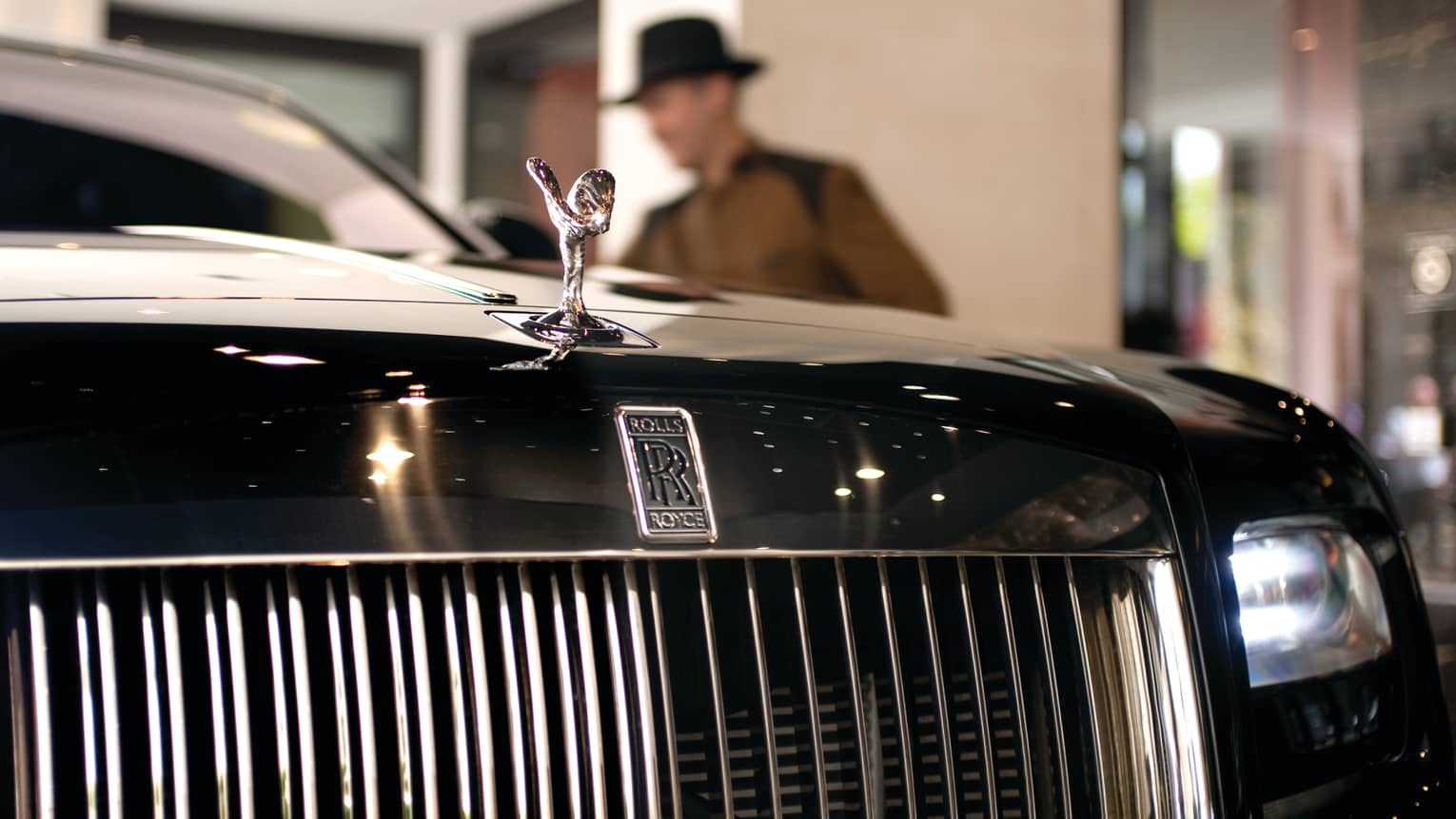 Close-up of Rolls Royce luxury car front grill, man in top hat in background