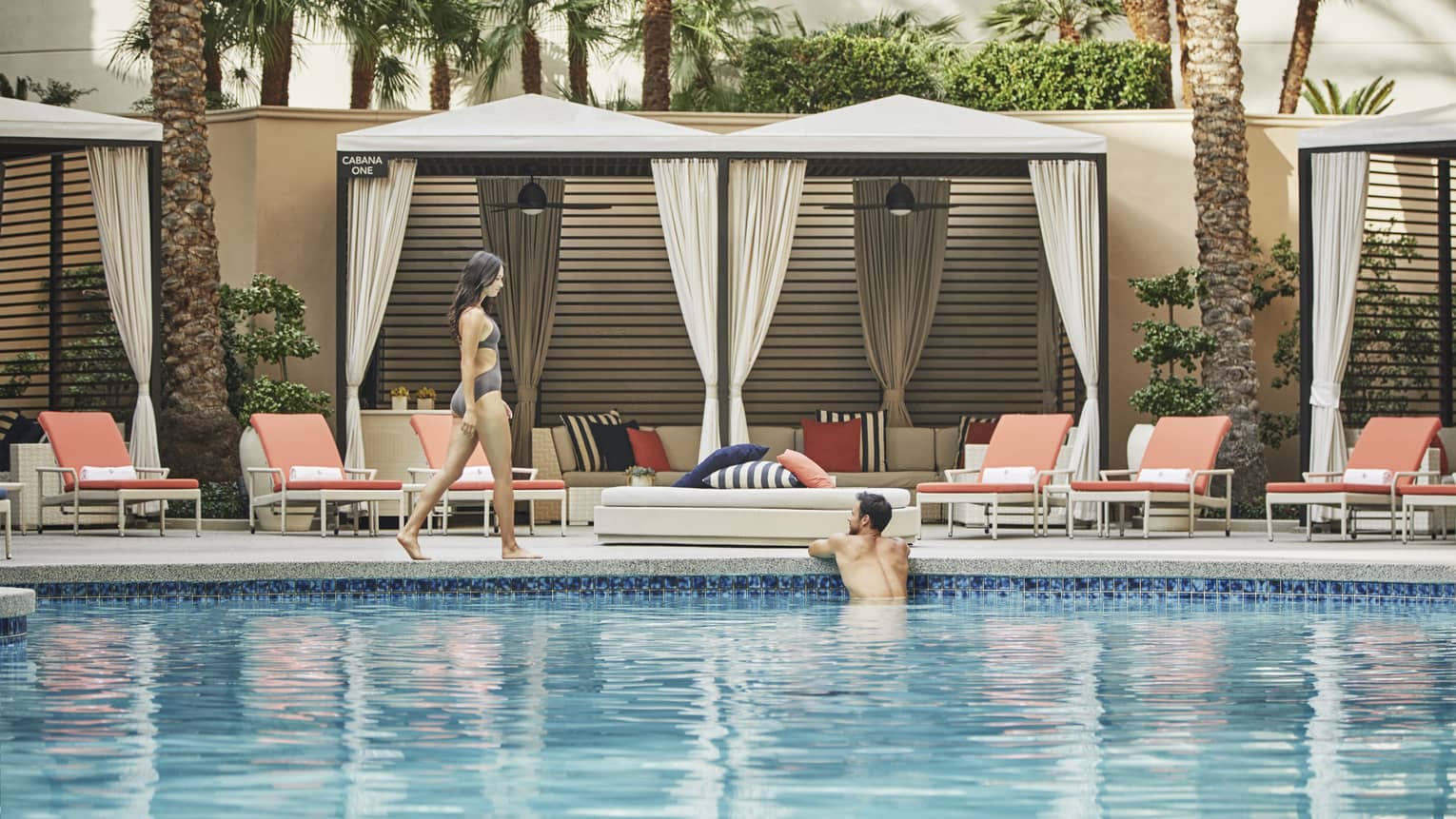 A woman walking next to a pool while a man is relaxing in it, lounge chairs are next to the pool.