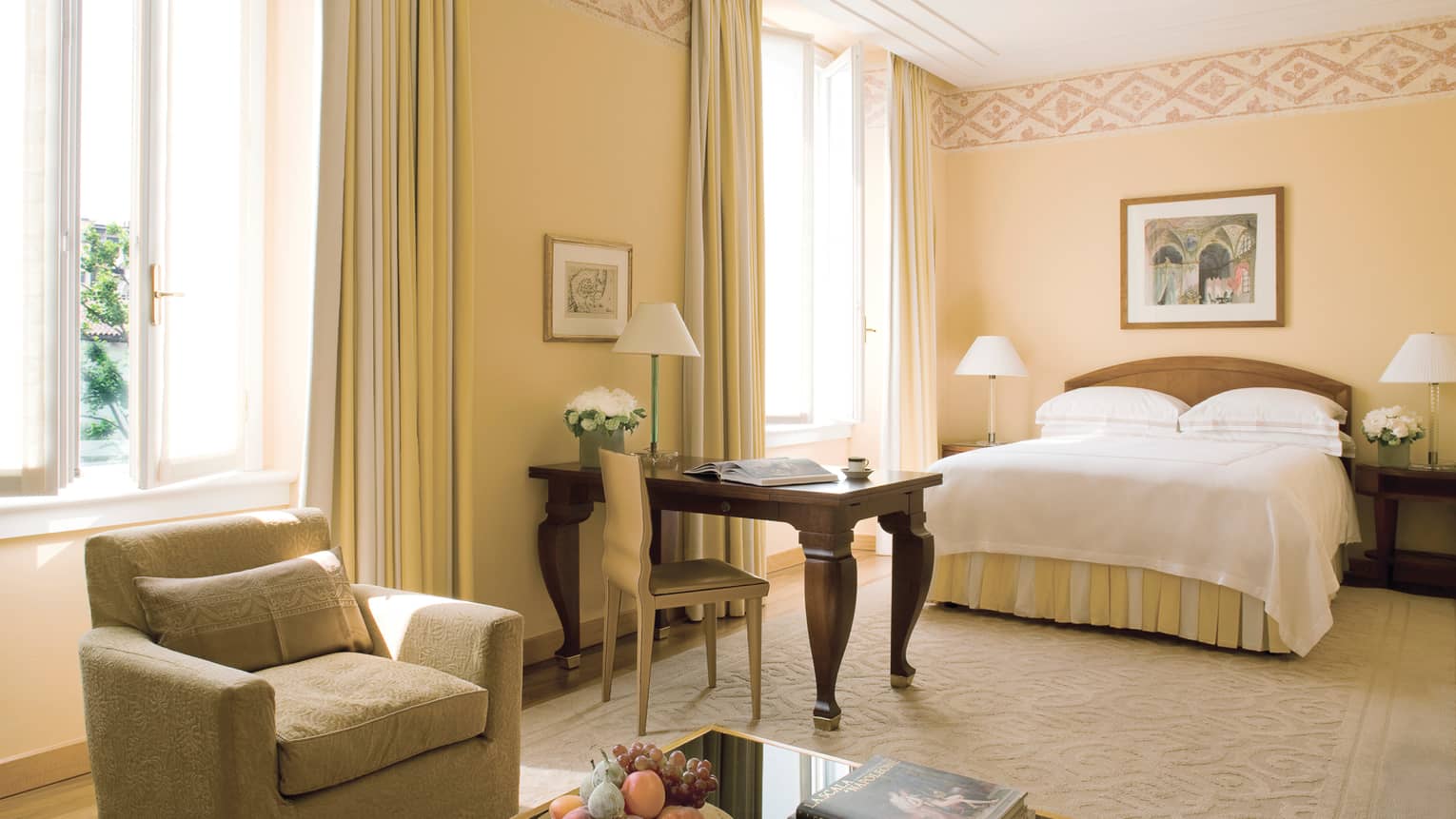 Deluxe Room armchair by table, fruit bowl, wood desk with chair, bed, sunny windows