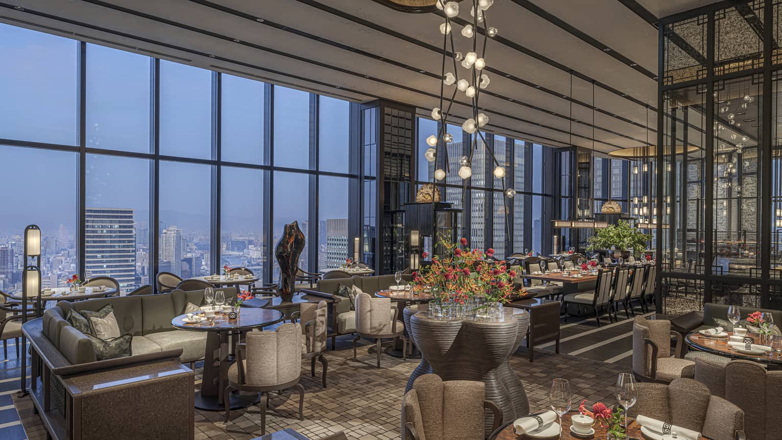 Expansive high-end restaurant warmly lit by modern chandeliers with a bank of windows offering aerial views of the city