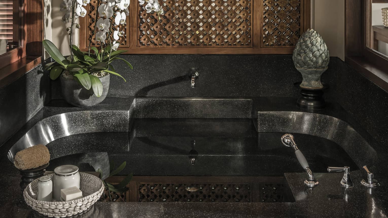 Black stone bathroom tub with potted plant and sculpture on ledge, basket with soaps and jars 