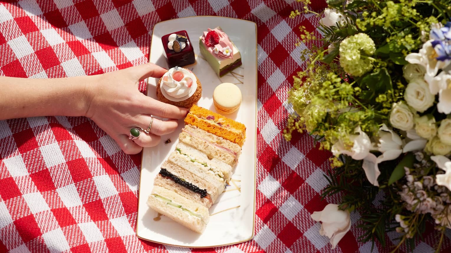 A hand reaches for a cherry-topped tart on a plate of dainty sandwiches and sweets on a red-and-white checked picnic blanket.