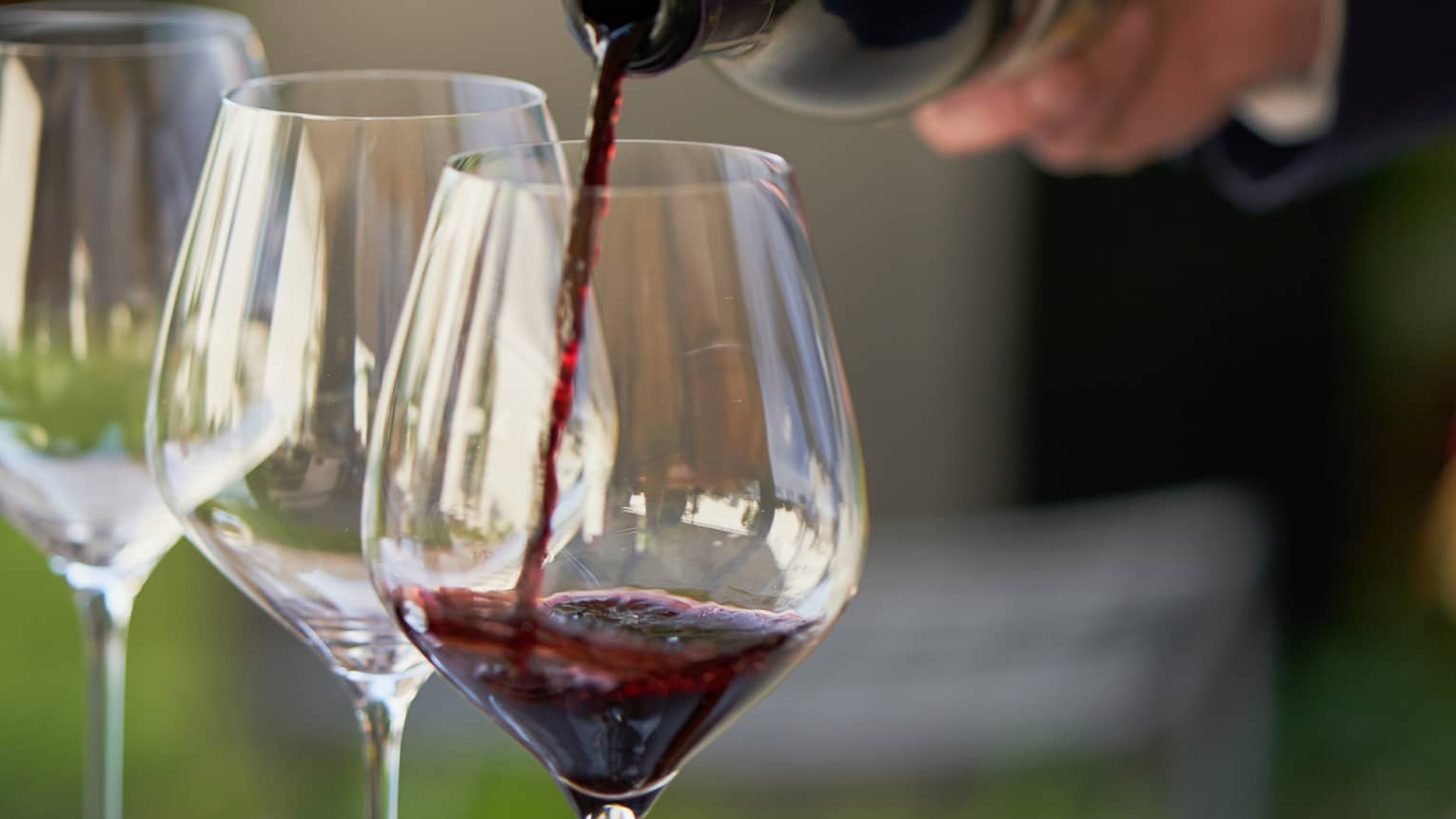 A close up of hands pouring a bottle of red wine into a clear wine glass that rests on a wooden tray, outdoors