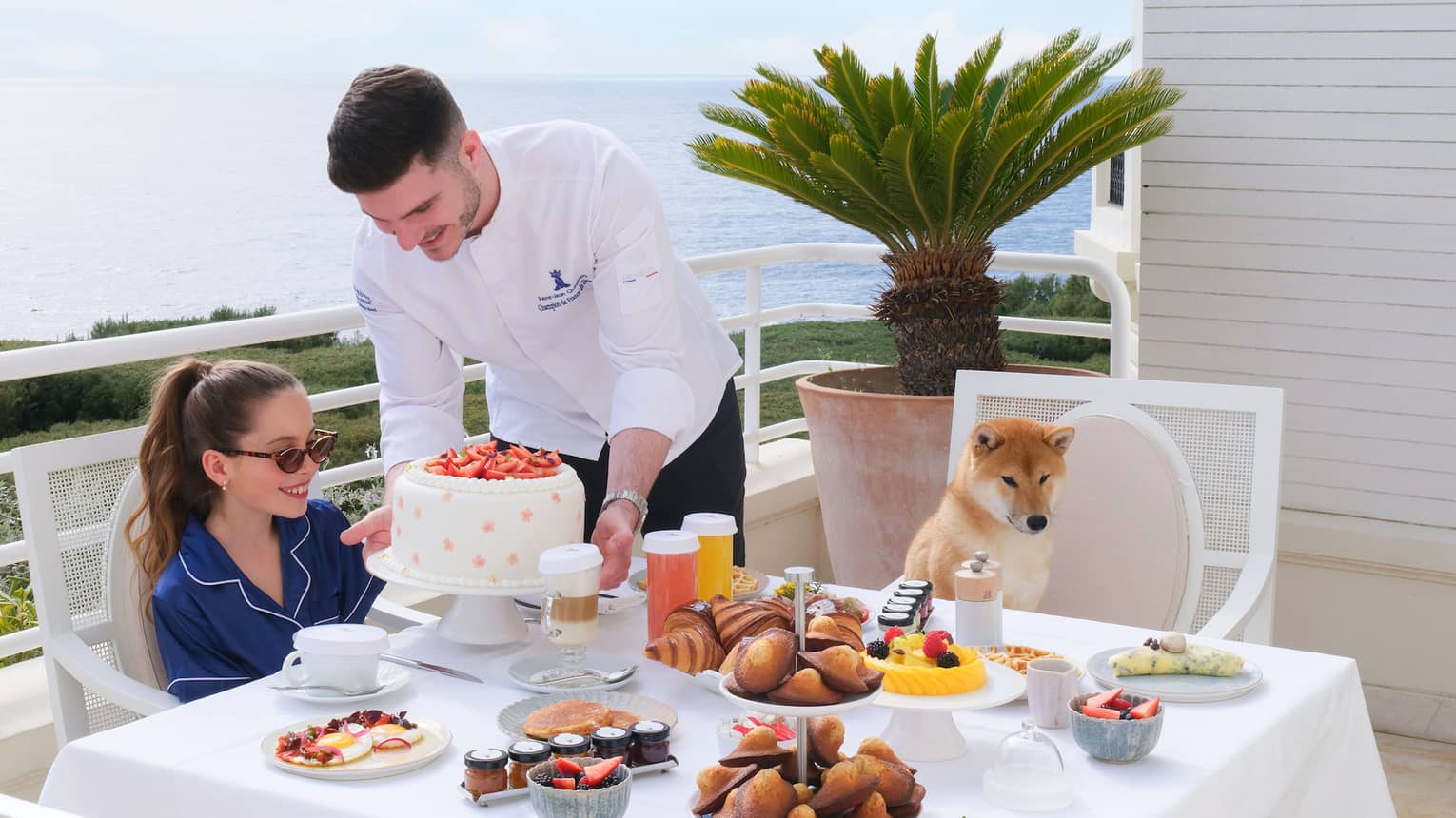 Chef presenting a white cake to child having breakfast with her dog at terrace table overlooking the sea