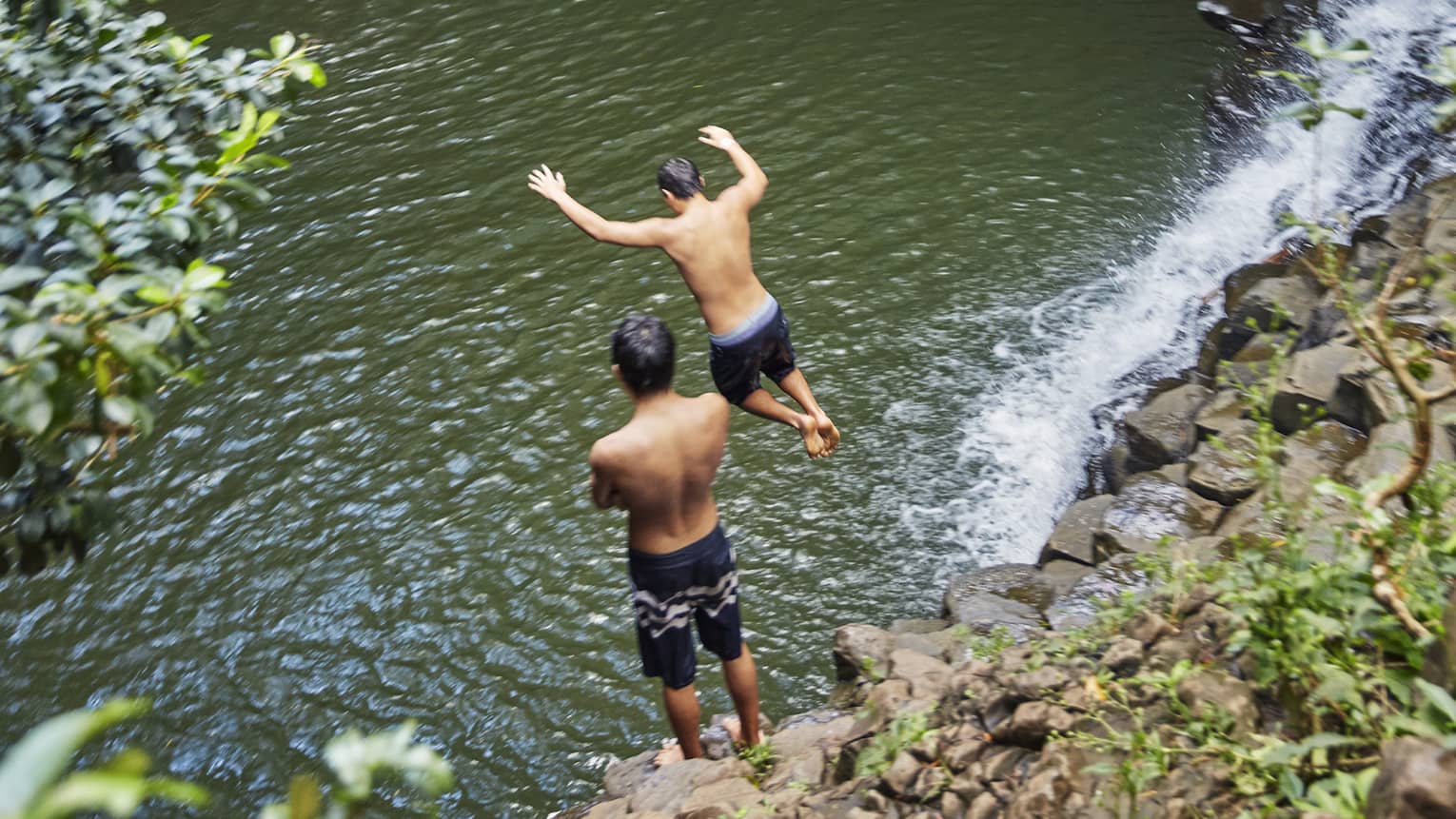 Two people leap from rocks to water below waterfall on the road to Hana