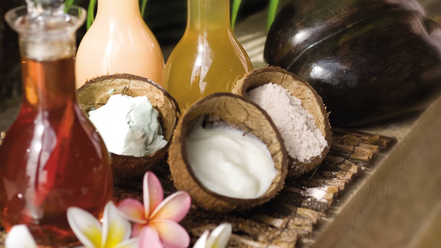 Three open coconut shells filled with white creams and lotions, glass bottles with infused oils, flowers